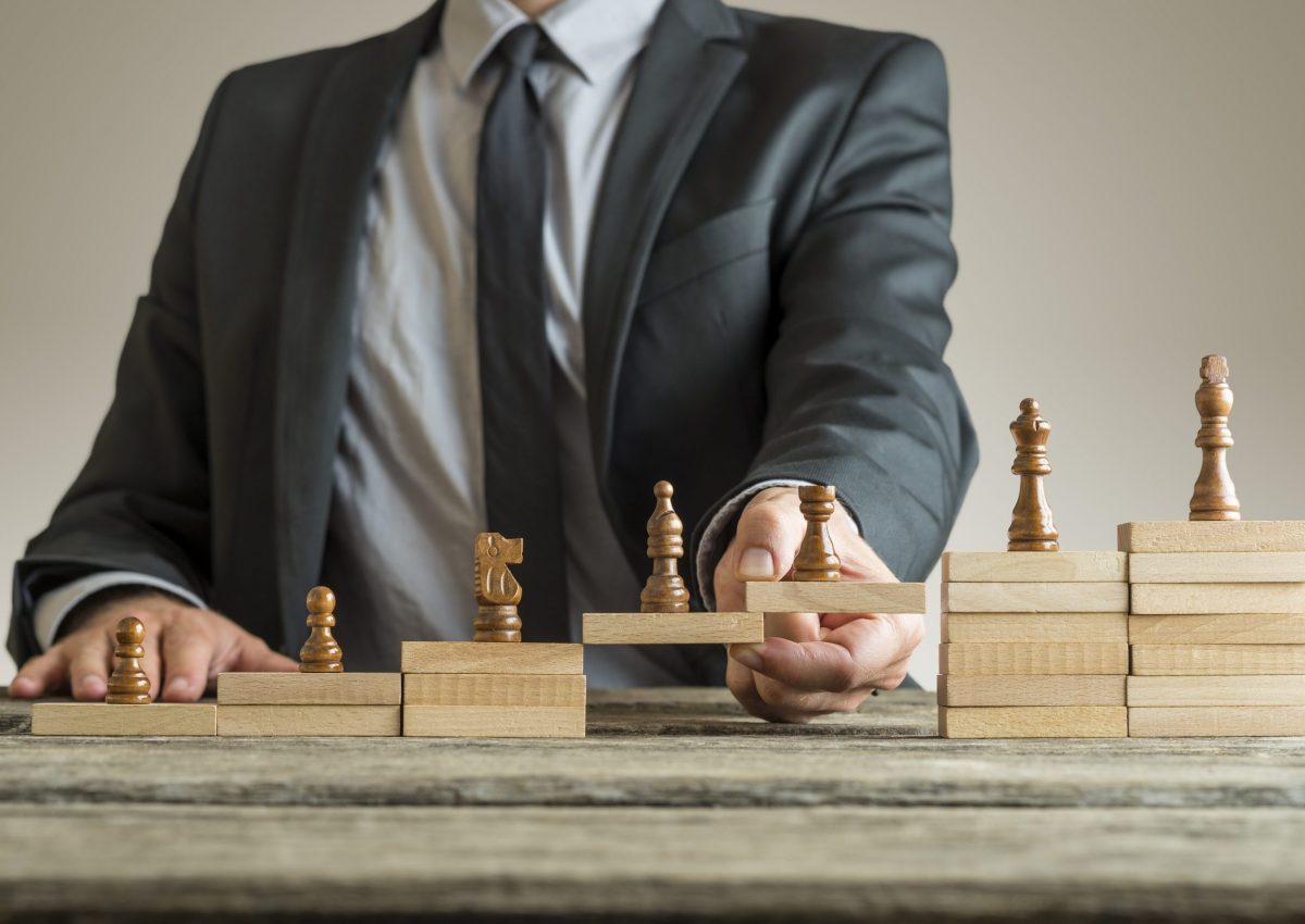 Conceptual image of career management with a businessman forming a bridge of wooden building blocks for chess pieces developing from pawn to king.